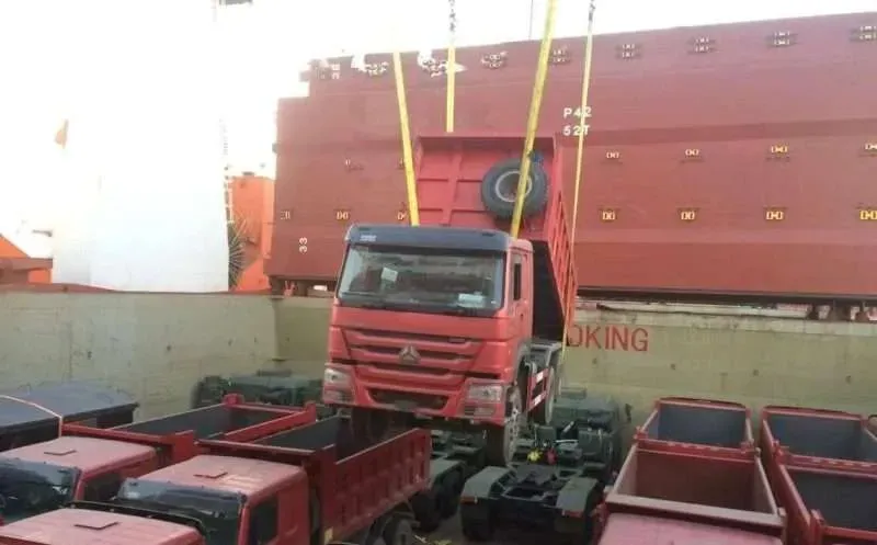 A red HOWO dump truck lifted by ship's crane during loading at the port