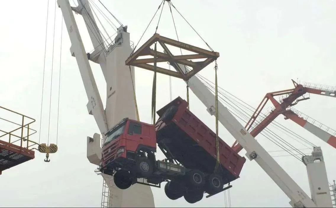 A red HOWO dump truck lifted by port cranes during loading