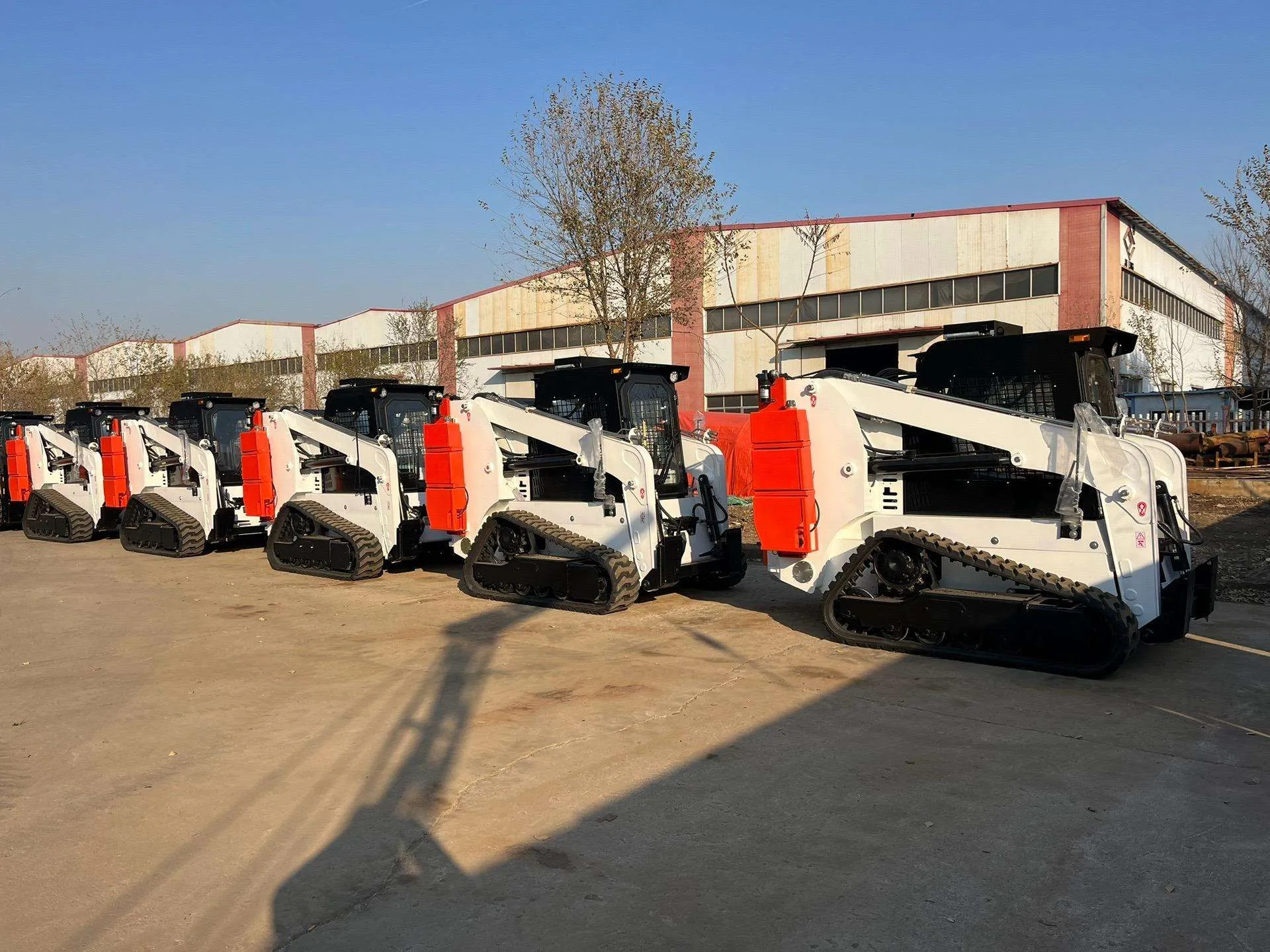 Six white and orange tracked skid steer loaders lined up in the factory yard
