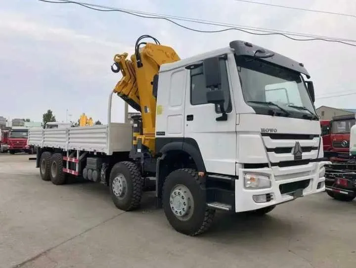 White 8x4 four-axle truck mounted crane with the yellow knuckle boom stowed over the cargo bed, front three-quarter view in the yard